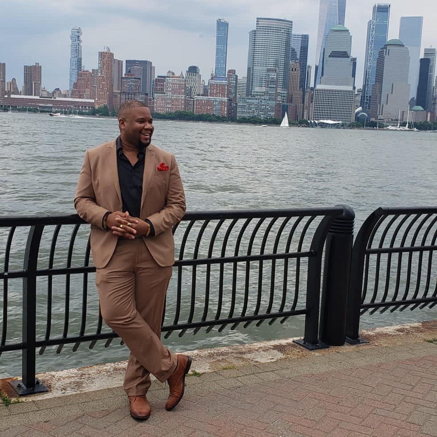 Jonathon Cummings in a tan suit on a waterfront promenade, the Lower Manhattan skyline behind him